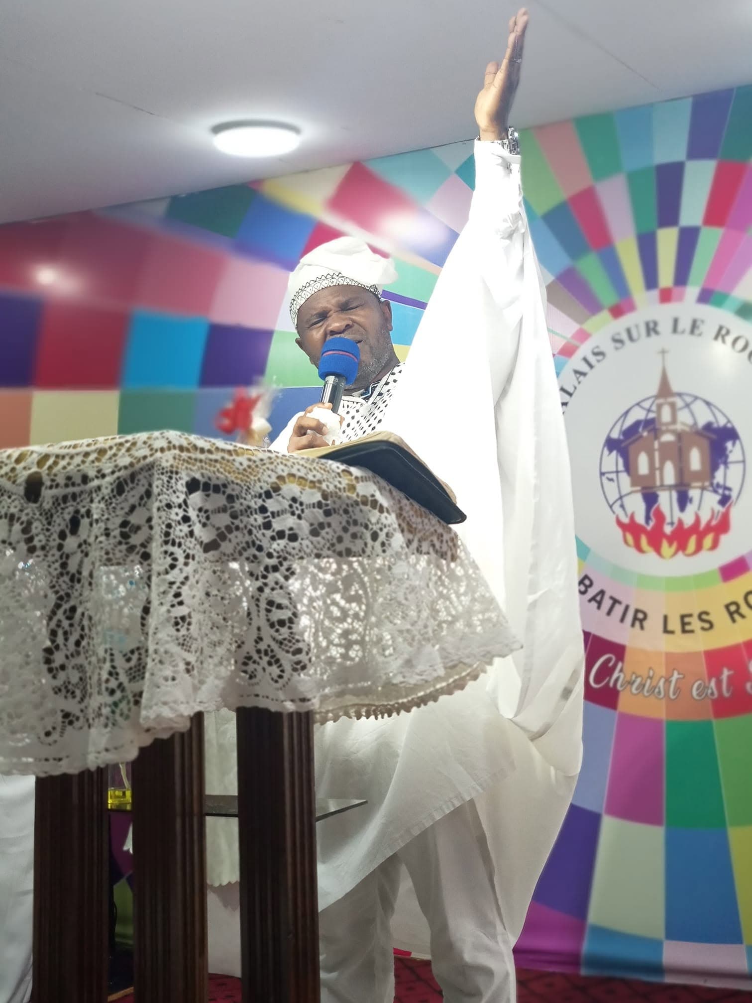 L'apôtre Daniel MBISSOKO au pupitre, bras levé, devant le logo de l'église